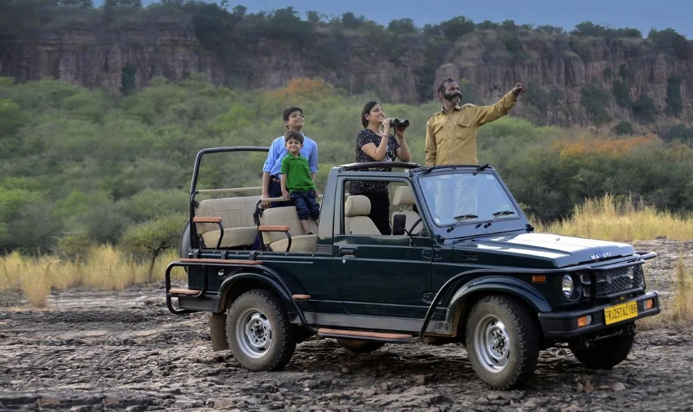 A family stands up in a safari jeep in Ranthambore National Park as their guide points out wildlife against a backdrop of forested cliffs